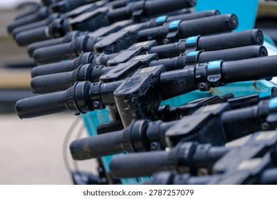 Close up view of multiple black rubber handles and brake levers of electric rental scooters parked orderly in a row on a city street.