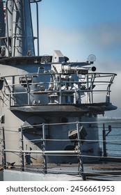 Close up view of naval ship superstructure with gun mount, radar dishes, and communication devices. Deck details in view, blurred water background. Likely in San Francisco. Sunny day.