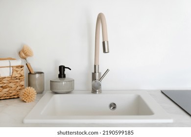 Close Up View Of New Faucet With A Water Mixer Above An Empty White Sink, Next To Dishwashing Accessory. Organic Detergent In Soap Dispenser Bottle, Bamboo Brush, Towel In Wicker Box On Kitchen Table