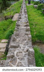 Close view of an old, irregular stone embankment or weir structure featuring rough hewn rocks set along the edge of a narrow river or canal with grassy banks, Lostwithiel, United Kingdom