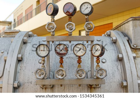 Close up view of old rusty damaged metal meter, valve tube system and gas or liquid tank for industry. Detail of broken circular measurement meter with rusty dial.