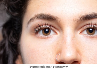 A close up view on the face of a young pretty Caucasian girl with brown eyes and long lashes. She looks towards the camera and has a small ring pierced in her nose.