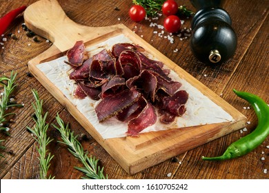 Close Up View On Tasty Sliced Beef Jerky On Parchment On A Wooden Board On A Wooden Background In A Composition With Spices. Beer Snack. Pastirma