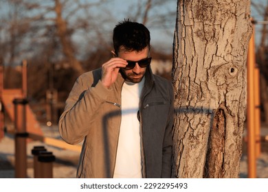 close up view on the upper half of the body of a young adult man, wearing casual, modern and fashion clothes and dark sunglasses, while being leant to a tree in a park, in the afternoon