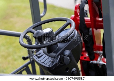 A close view of the operator control console on a compact articulated loader shows an ergonomic steering wheel and integrated control buttons mounted on a metal panel. Surrounding structural components and red machine elements indicate the front section of the loader used for agricultural, construction, and municipal work.
