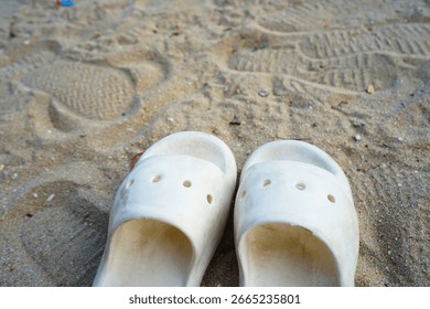 Close up view of a pair of white plastic sandals on a sandy beach, footprints visible in the sand.