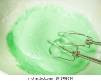 Close up view of pastel or mint green colored frosting and metal beaters in a white ceramic mixing bowl sitting on top of a kitchen counter. - Shutterstock ID 1042909834