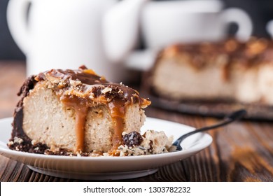 Close Up View Of A Piece Of Peanut Butter Chocolate Caramel Cheesecake Pie And Cup Of Coffee On Wooden Background With A Coffee Pot And Cheesecake In The Background. A Sweet Coffee Break Concept 