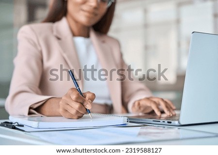 Close up view of professional busy African American business woman manager executive working on laptop computer writing notes in notebook at work elearning in corporate office.