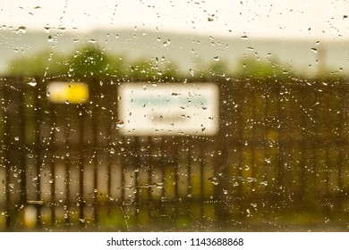 Close Up View Of Raindrops On A Train Window With A Station Nameplate Blurred In The Background.