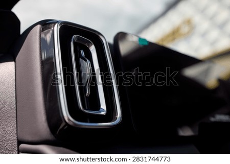 Close view of rectangular car interior air vent with chrome trim, glossy black grille, and textured matte dashboard surface, lit by soft daylight that reveals clean metallic detail and modern cabin
