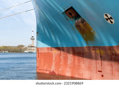 Close up view of the red and blue colored bow a ship with anchor hole, Load line mark and with patchy paintwork with in background the Maas river in Rotterdam, the Netherlands