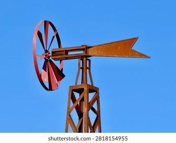Close view of red painted windmill rotor and rusted metal tail on tall lattice tower under clear blue sky, with weathered surface, bolted hub, curved blade shape, and rugged rural character