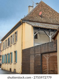 Close view of a residential building facade with pale yellow walls and blue wooden shutters aligned along multiple windows. The structure features a sloped tiled roof with a satellite dish and antenna, alongside a modern wooden fence in the foreground. The combination of traditional architecture and contemporary elements stands out under a soft cloudy sky in a quiet street setting.