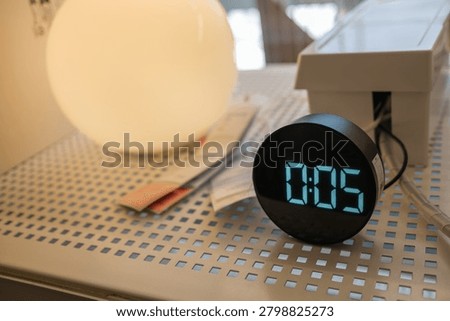 Close up view of a round digital clock showing time on a white perforated shelf next to a glowing spherical lamp.