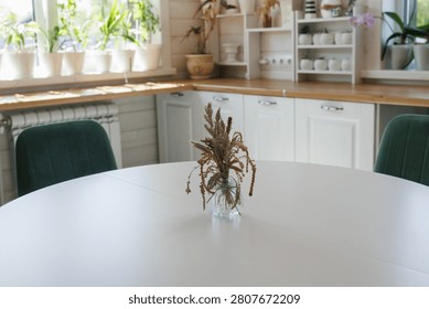 Close view of round white dining table featuring small glass vase with dried brown grasses at center, set inside bright modern farmhouse kitchen with wooden countertop, white cabinetry, potted plants