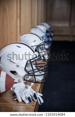 Close up view of a row of American football helmets sitting in a locker room before an football game. Gloves and a football also sitting on the bench in the lockerroom

