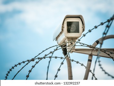 Close up view of security camera hanging among barbwire in prison or other guarded object with blue sky background. Modern ways of supervision. Using new technology in security and safety.