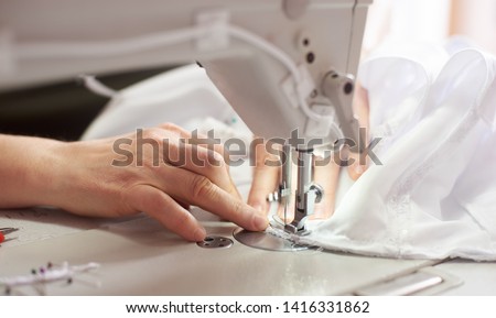 Close up view of sewing process. Female hands stitching white fabric on professional manufacturing machine at workplace. Seamstress hands holding textile for dress production. Light blurred background