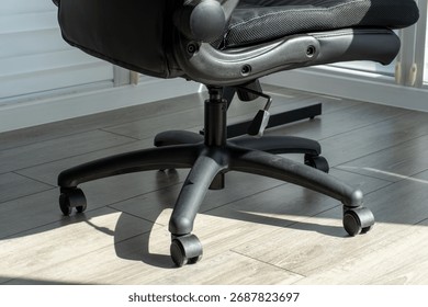 A close up view showcasing the lower section of a contemporary black adjustable office chair. The robust five-star base with smoothly rolling casters sits on a pale wood grain laminate floor. 