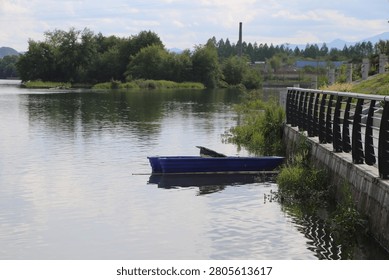 A close view shows two long open boats resting in shadow near a paved riverside edge. Oars, seats, and worn surfaces are visible inside, while the dark water reflects the boats and railing.