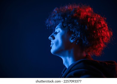 Close up view from the side. Young man with curly hair is indoors illuminated by neon lighting.