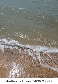 Close Up View Of Small Ocean Waves Breaking On Pebbled Shoreline With Foamy Water And Wet Sand Texture