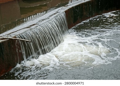 Close up view of a small silky cascade of water flowing over a man-made concrete wall and splashing into a pool of water below.