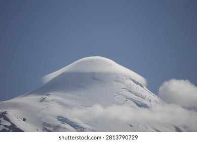 Close view of the snow covered summit of Osorno Volcano wrapped by a smooth lenticular cloud under clear blue sky.