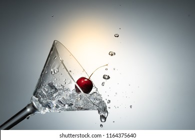 Close up view of splash water with falling cherry in a martini glass among ice in white background with flare .