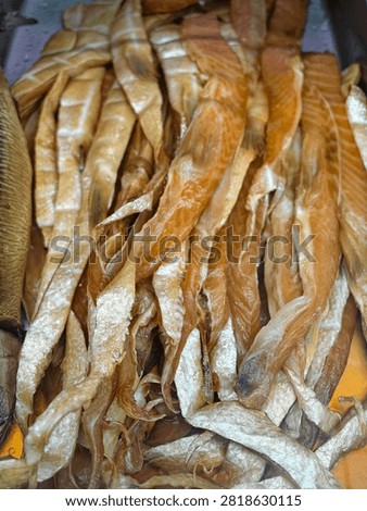 Close view of stacked dried fish strips with glossy golden brown tones, shiny texture, and layered shapes inside indoor market display, suggesting food storage, trade, and traditional preservation