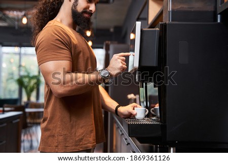Close up view of the startup coworker man making coffee during office break time while standing near the coffee machine and pushing at the button with his finger