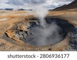 Close view of a steaming mud pot at Hverir geothermal area, Namaskard, North Iceland, cracked sulfur-crusted earth and billowing steam against distant mountains.