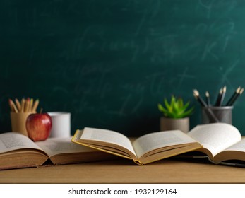 Close Up View Of Study Table In Classroom With Opened Books, Stationery And Apple In With Blackboard Background