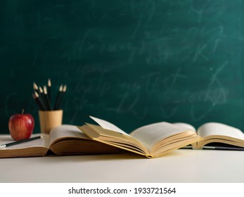 Close up view of study table with opened books, pencils and apple on white desk with chalkboard wall background