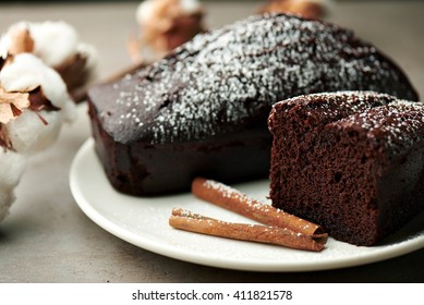 Close View At Tasty Chocolate Cake On A White Plate, With Ceramic Cup, Cinnamon Sticks And Cotton Flowers On A Grey Background, Sunday Morning Breakfast.