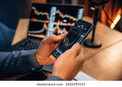 Close up view of trader's hands that holding phone with crypto data, laptop on the table.