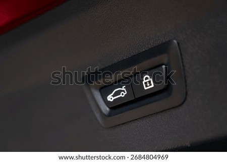 Close up view of two buttons on a interior panel, featuring a car trunk icon and a lock symbol. Red trim is visible in the background.