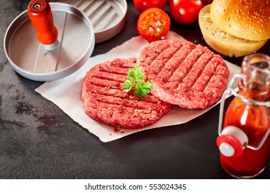 Close Up View Of Two Raw Beef Hamburger Patties Topped With Sprig Of Fresh Parsley Resting On Square Of Wax Paper And Surrounded By Buns, Press And Fresh Ingredients With Copy Space In Foreground