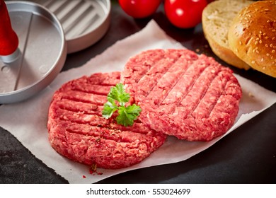 Close Up View Of Two Raw Beef Hamburger Patties Topped With Sprig Of Fresh Parsley Resting On Square Of Wax Paper And Surrounded By Buns, Press And Fresh Ingredients