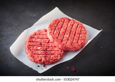 Close Up View Of Two Raw Beef Hamburger Patties On Square Of Wax Paper Resting On Dark Gray Stone Textured Counter Surface With Copy Space