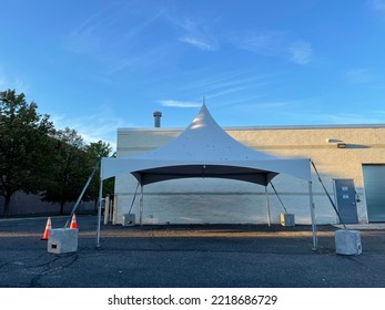 The Close Up View Of A White Tent Set Up Behind A Building. Each Corner Of The Tent Is Weight Down With Heavy, Cement Blocks.