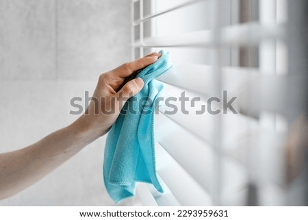 Close up view of woman cleaning white window blinds with a blue microfibra rag indoors. Housework and housekeeper concepts