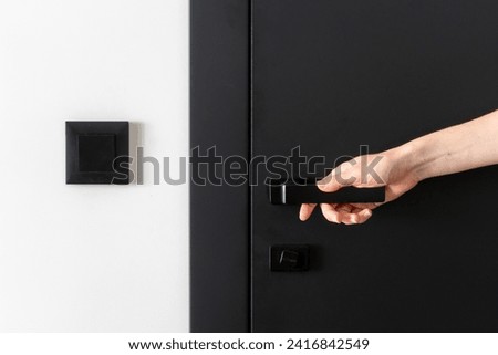 Close up view of woman hand closed modern black door with metal handle and lock. White wall with light switch near the entrance. Privacy and safety concept. Copy space