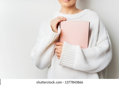 Close view of woman in white woolen sweater holding a book with empty pink cover in hands. Free space for your mock up of reading book concept background.
