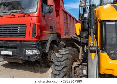 Close view of yellow loader beside red dump truck in industrial yard under blue sky, showing big tires, metal arm, cab details, and rough worksite conditions for transport, construction, and