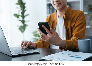 Close up view of young Asian businessman looking at smartphone screen while typing on laptop computer. - Shutterstock ID 2298048099