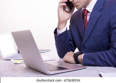 Close up view of a young businessman working on his desk white background. Two computers on the table. Man making a phone call while looking at laptop screens. Red tie and blue suit. - Powered by Shutterstock