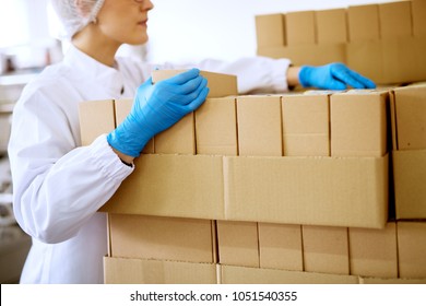 Close Up View Of A Young Focused Female Worker In Sterile Cloths And Blue Rubber Gloves Counting Stacked Boxes In A Factory Storage Room.