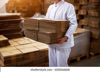 Close up view of a young hard working employee in sterile cloths carrying the stack of brown cardboard folded boxes from the factory cargo room.
